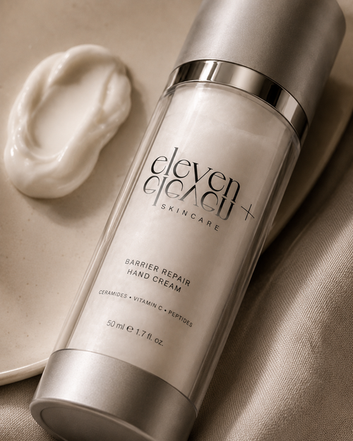 A bottle of Eleven Eleven+ Barrier Repair Hand Cream with a silver cap and clear body, shown on a beige fabric surface next to a dollop of white cream.