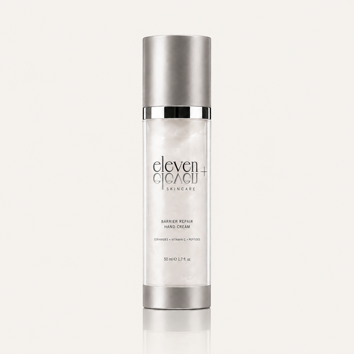 A tall cylindrical bottle of Eleven Eleven Skincare Barrier Repair Hand Cream, featuring a metallic silver cap and base with a white body, set against a light beige background with a soft reflection.